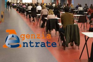 Bando Agenzia delle entrate: persone sedute in aula che compilano il bando. Logo Agenzia Entrate
