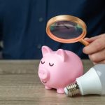 Uomo con lente d'ingrandimento su salvadanaio e lampadina vicino: controllo dei consumi