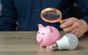 Uomo con lente d'ingrandimento su salvadanaio e lampadina vicino: controllo dei consumi