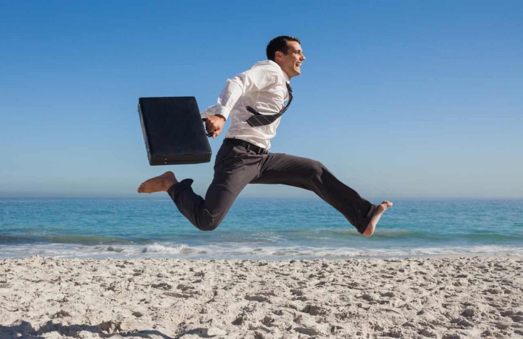 Lavoratore in camicia e cravatta che salta felice in spiaggia: ferie estive