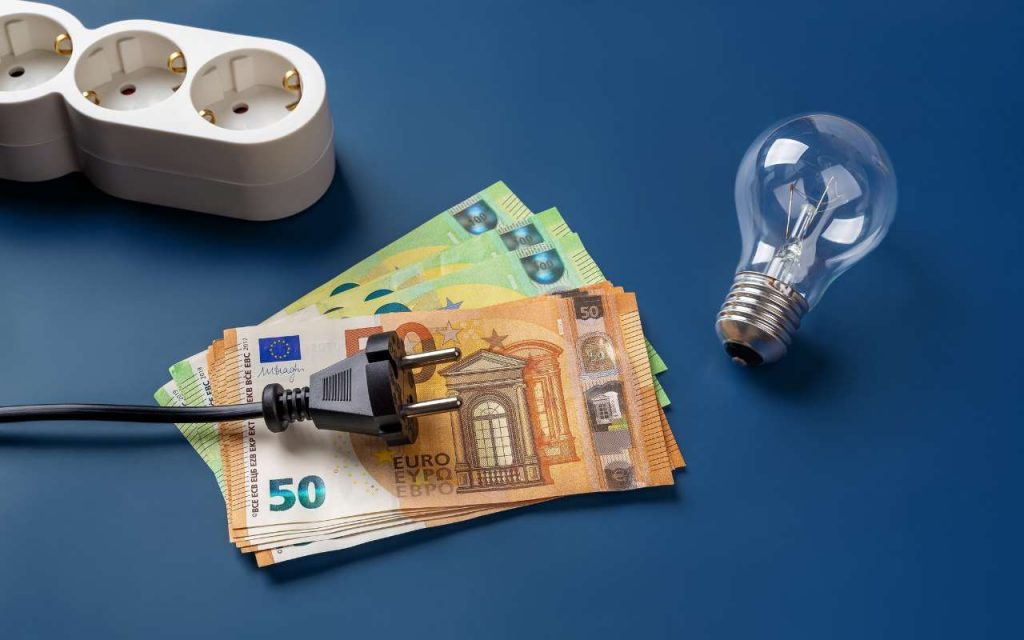 Banconote da 50 e 100€ sul tavolo, una spina della corrente, una lampadina e una ciabatta: spesa della luce